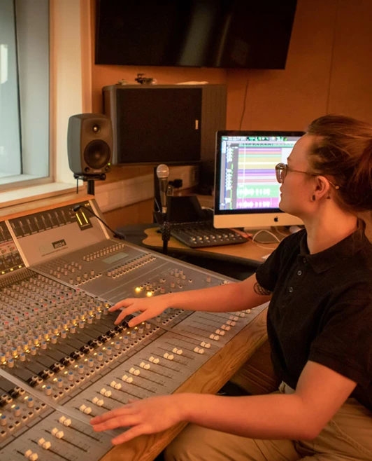 A student in a black polo shirt adjusts the controls on a mixing desk while monitoring a guitarist in a soundproofed studio. A student in a black polo shirt adjusts the controls on a mixing desk while monitoring a guitarist in a soundproofed studio.